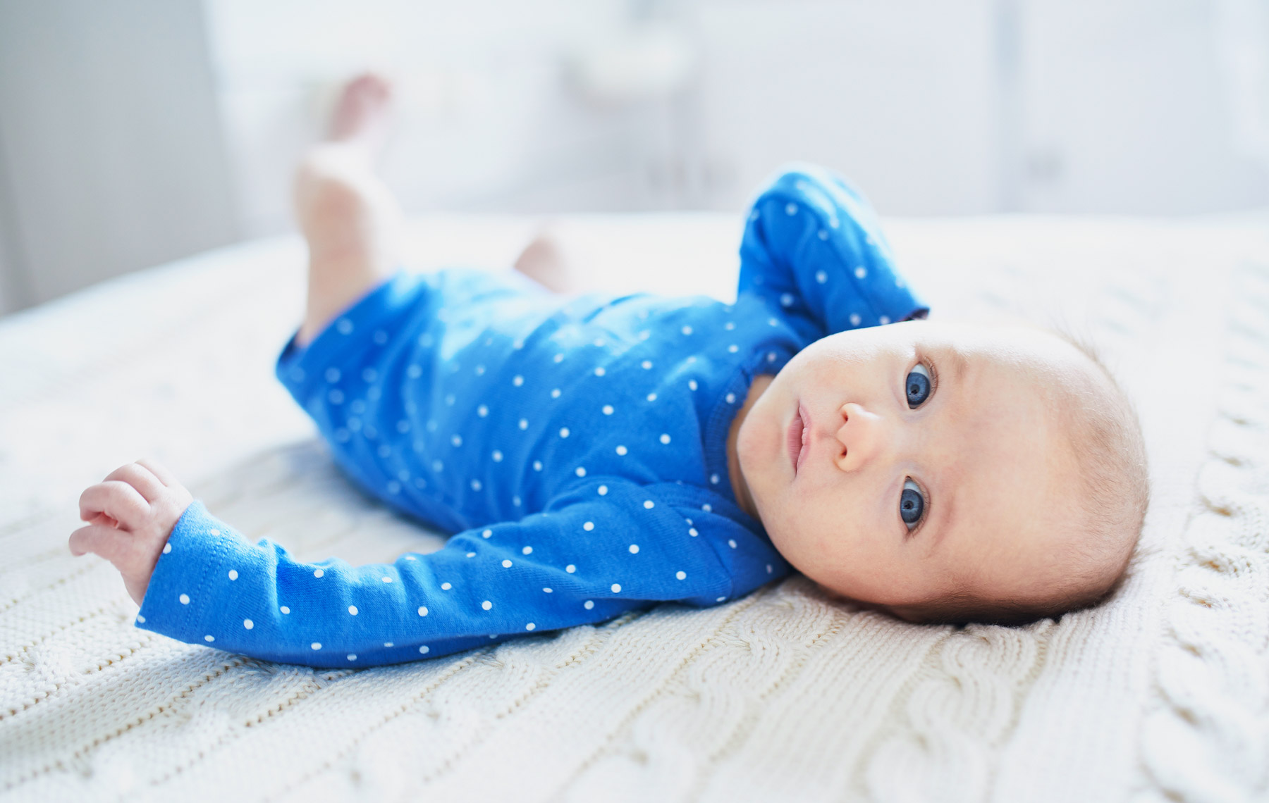 Ein Säugling in einem blauem Strampler liegt auf einer Decke und schaut den Betrachter mit blauen Augen an.