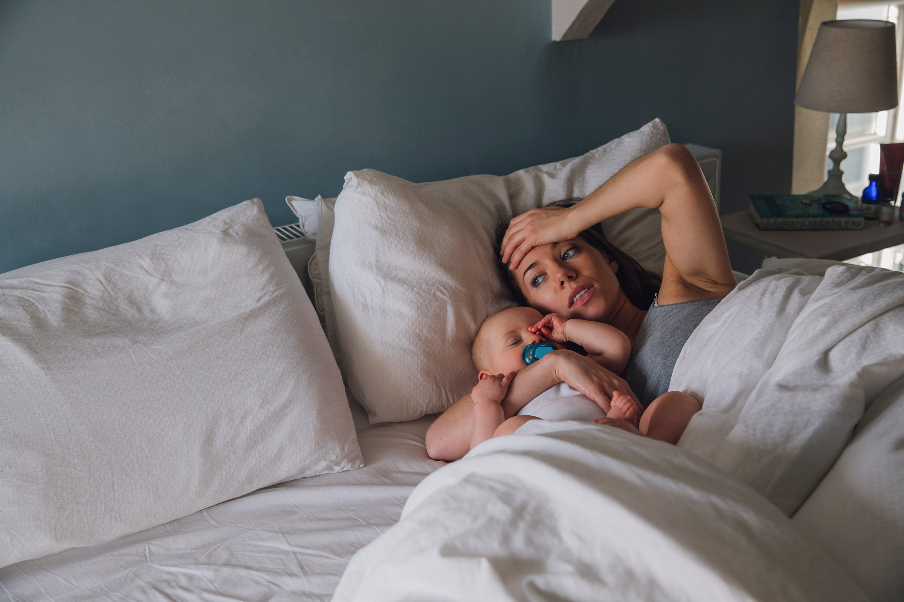 Eine Frau liegt mit ihrem Säugling im Bett. Sie hält sich die Hand auf die Stirn und macht einen überforderten Eindruck.