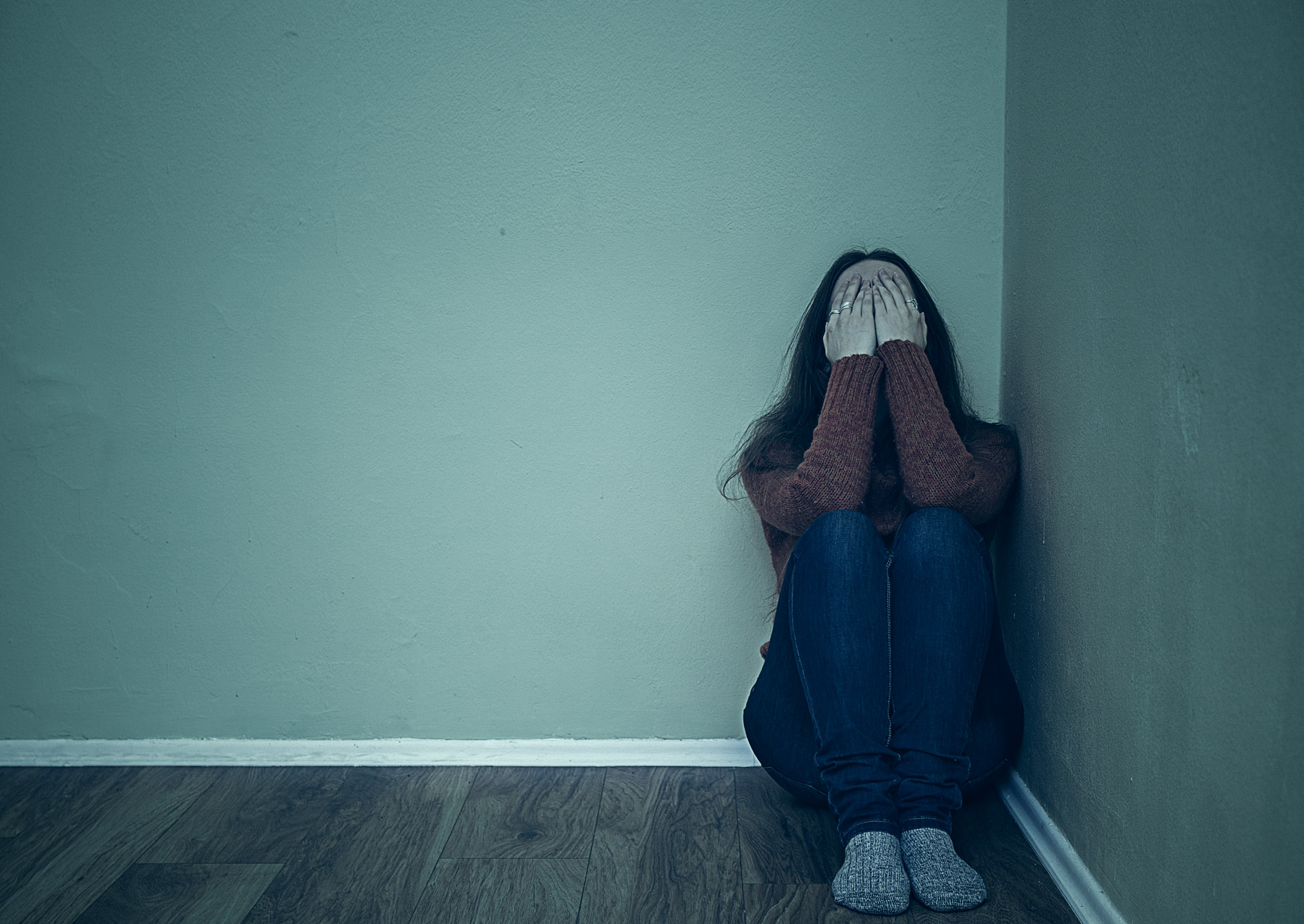 Eine Frau sitzt mit angezogenen Beinen in einer Raumecke. Sie hat die Hände vor dem Gesicht und wirkt verzweifelt.