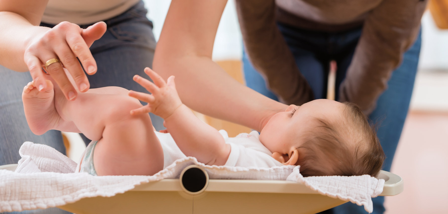 Ein Säugling auf einer Babywaage. Zwei Personen im Hintergrund von denen nur Beine und Hände zu sehen sind.