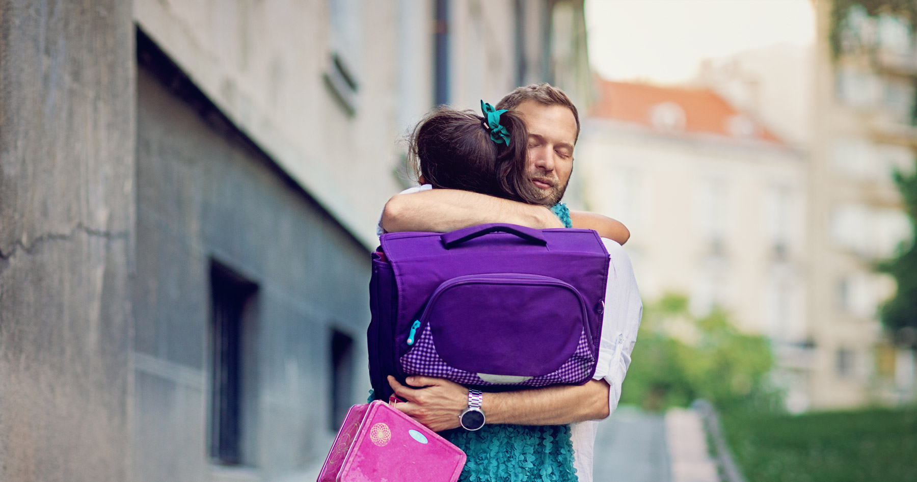 Ein Mann umarmt ein Mädchen mit einem Schulranzen. Das Mädchen ist von hinten zu sehen.