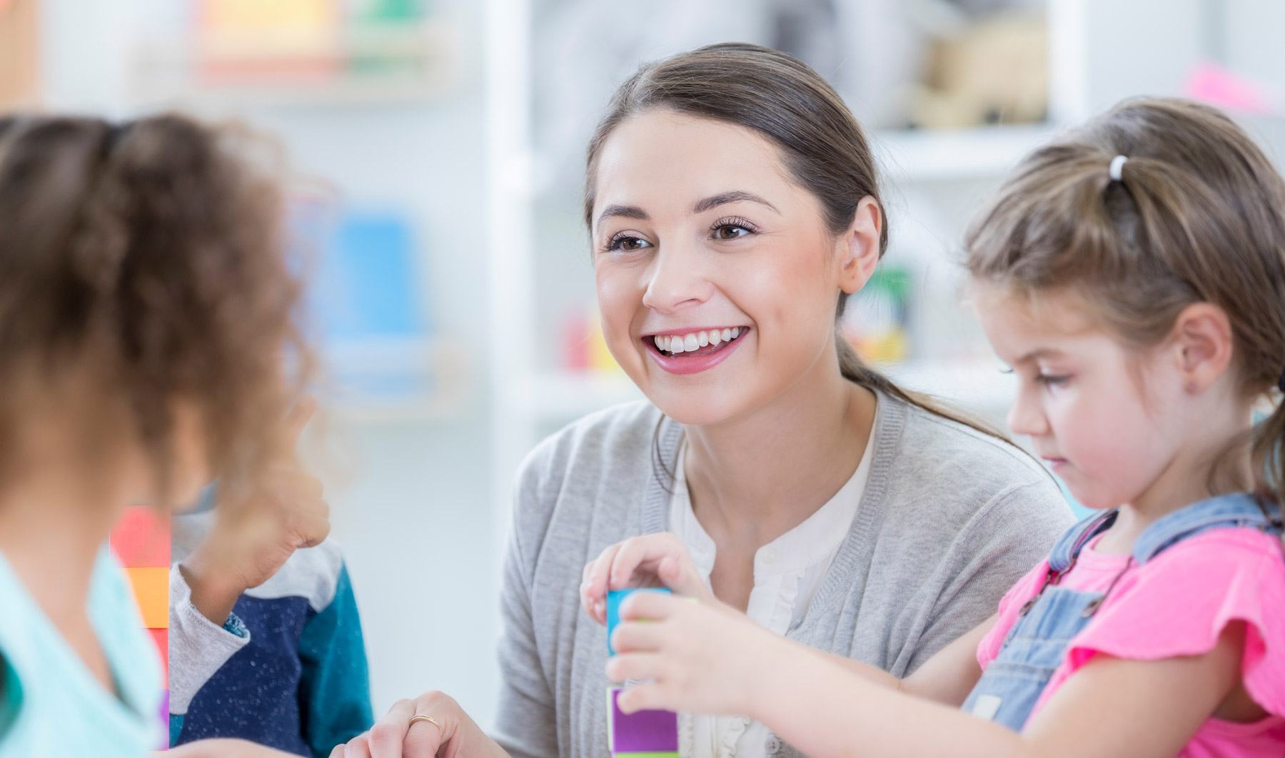 Kindertagespflege Eine junge Frau sitzt zwischen Kleinkindern, sie lächelt, die Kinder spielen.