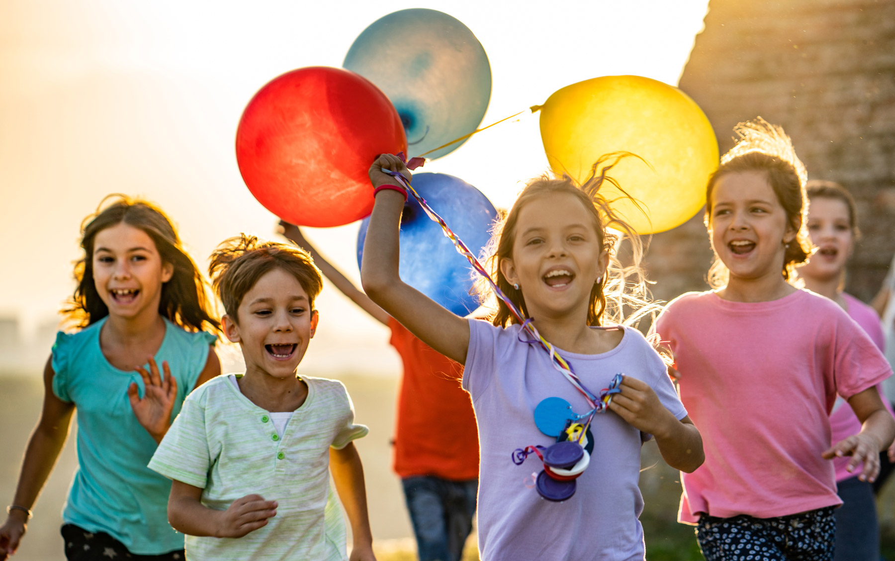 Eine Gruppe Kinder rennt dem Betrachter mit bunten Luftballons entgegen.