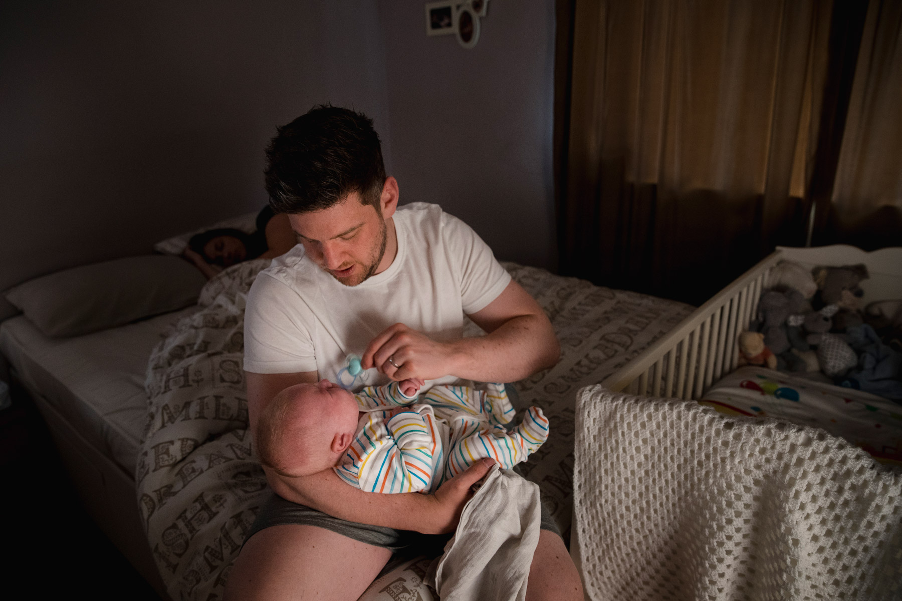 Ein junger Vater sitzt mit seinem Säugling auf der Bettkante. Im Hintergrund schläft die Mutter, das Baby weint.