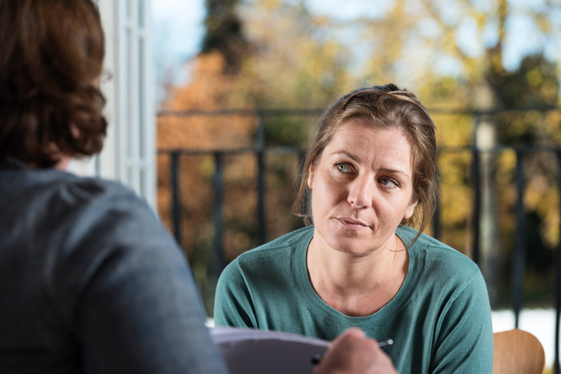 Zwei Frauen sitzen sich gegenüber. Eine Frau ist unscharf dargestellt und mit dem Rücken zum Betrachter platziert. Die zweite Frau sieht nachdenklich und angestrengt aus.