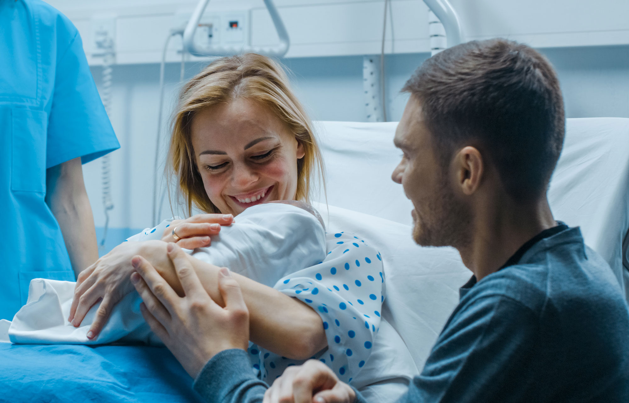 Eine Frau hält ihr Neugeborenes in den Armen, der Partner sitzt neben ihr. Sie befinden sich in einem Krankenhauszimmer.